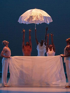 Photo of a theater stage with actors holding a white sheet and a large white umbrella.