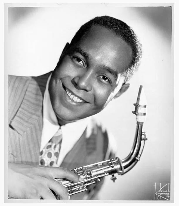 B/W portrait of a gentleman holding a saxophone and smiling for the camera.