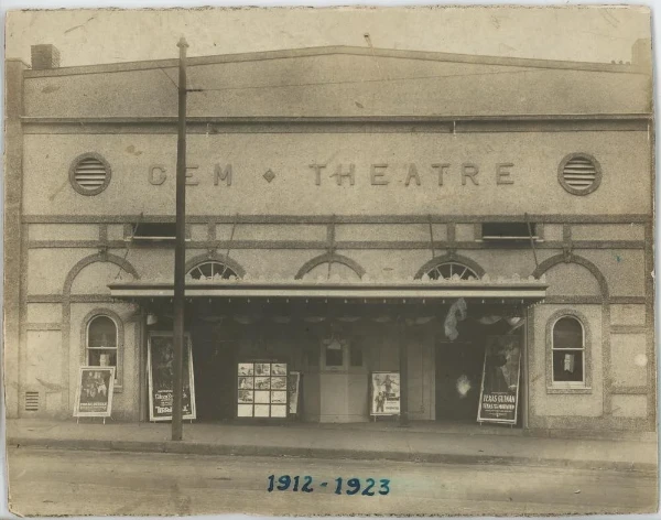 Sepia toned picture of a building with Gem Theater in large letters and standing advertisement signs along the sidewalk. 1912 - 1923 is written across the bottom in blue ink.