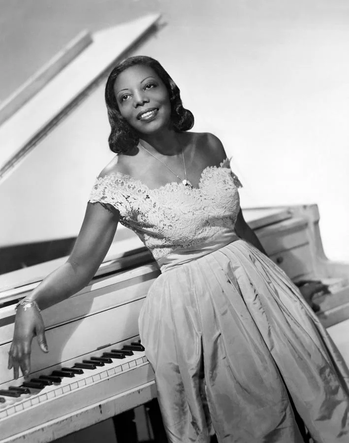 B/W photo of a woman smiling in a white dress in front of a white piano, one arm draped across the top, the other on the keys