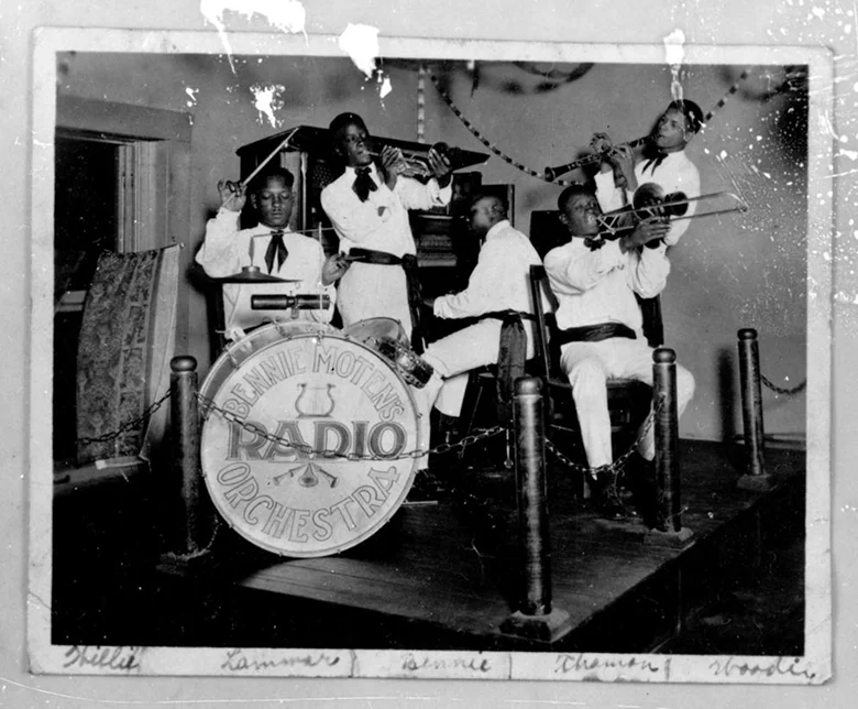 B/W photo of a five man orchestral band playing behind pylons. The men are all in white. We see a trumpet player, a trombone player, a clarinet player and a piano player. They're led by man behind a drum kit that reads 